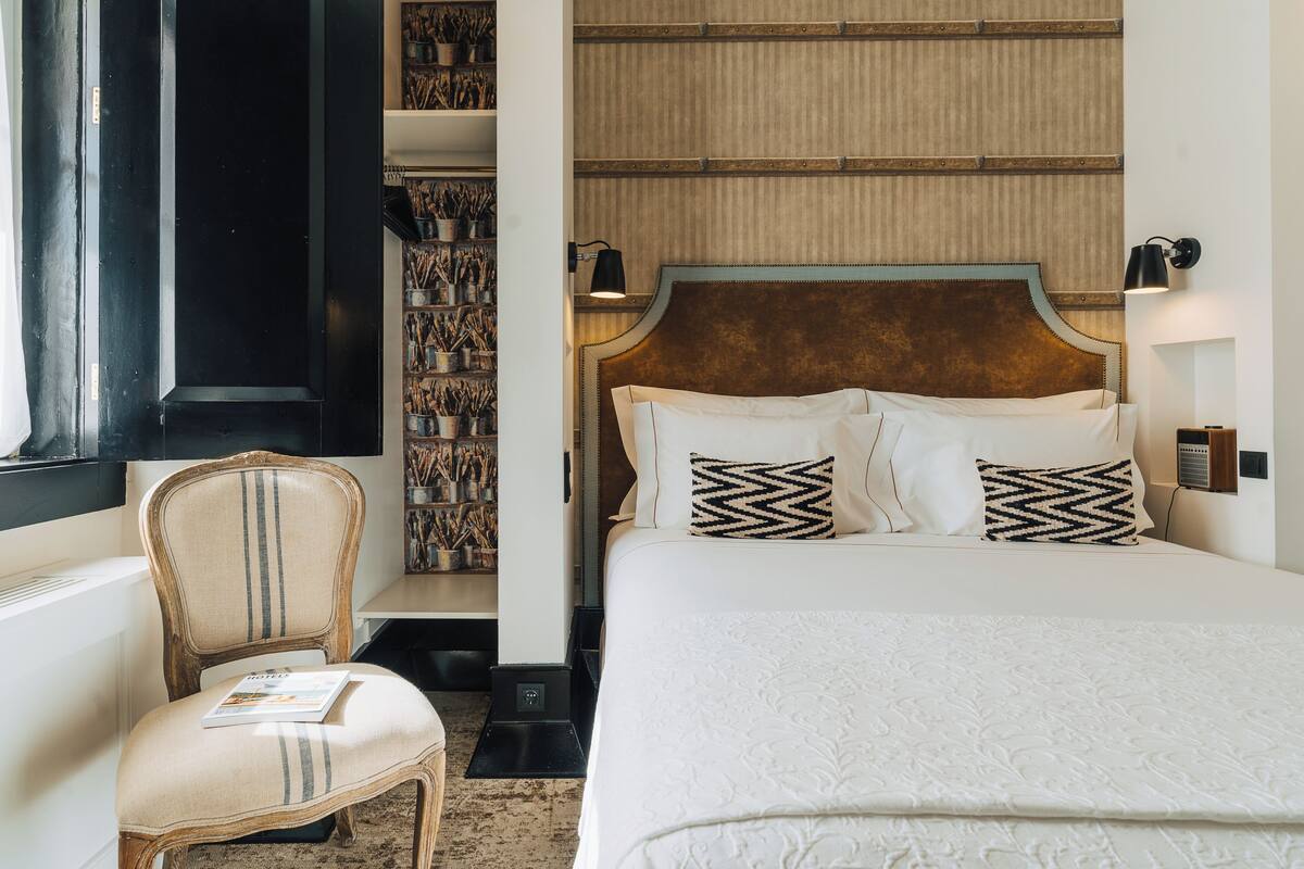 A hotel room with a large bed, a chair, a bookshelf, and a wall-mounted lamp.