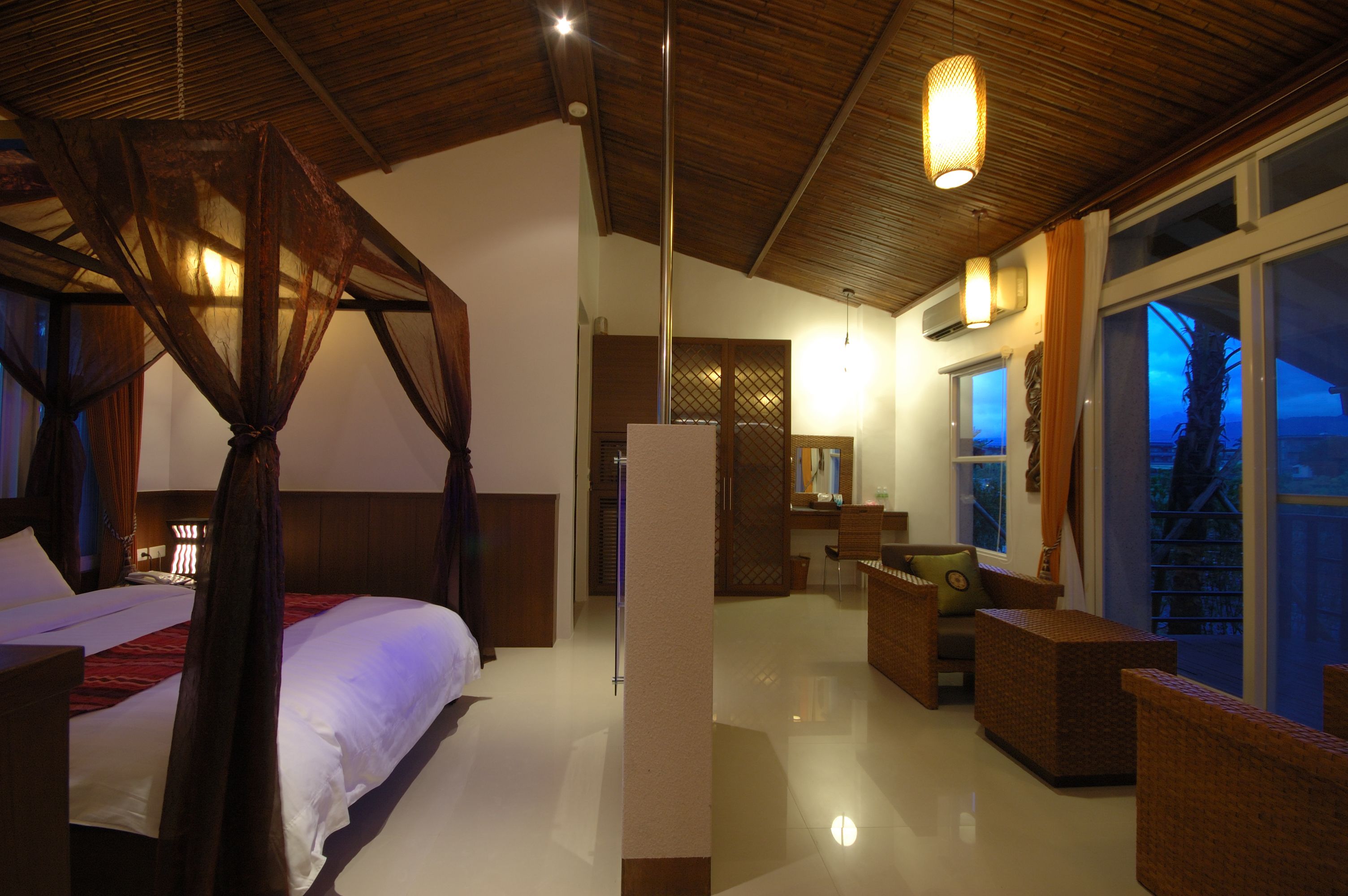 Un dormitorio espacioso con cama con dosel, una zona de estar con mobiliario de mimbre y un ventanal que ofrece vistas al exterior.
