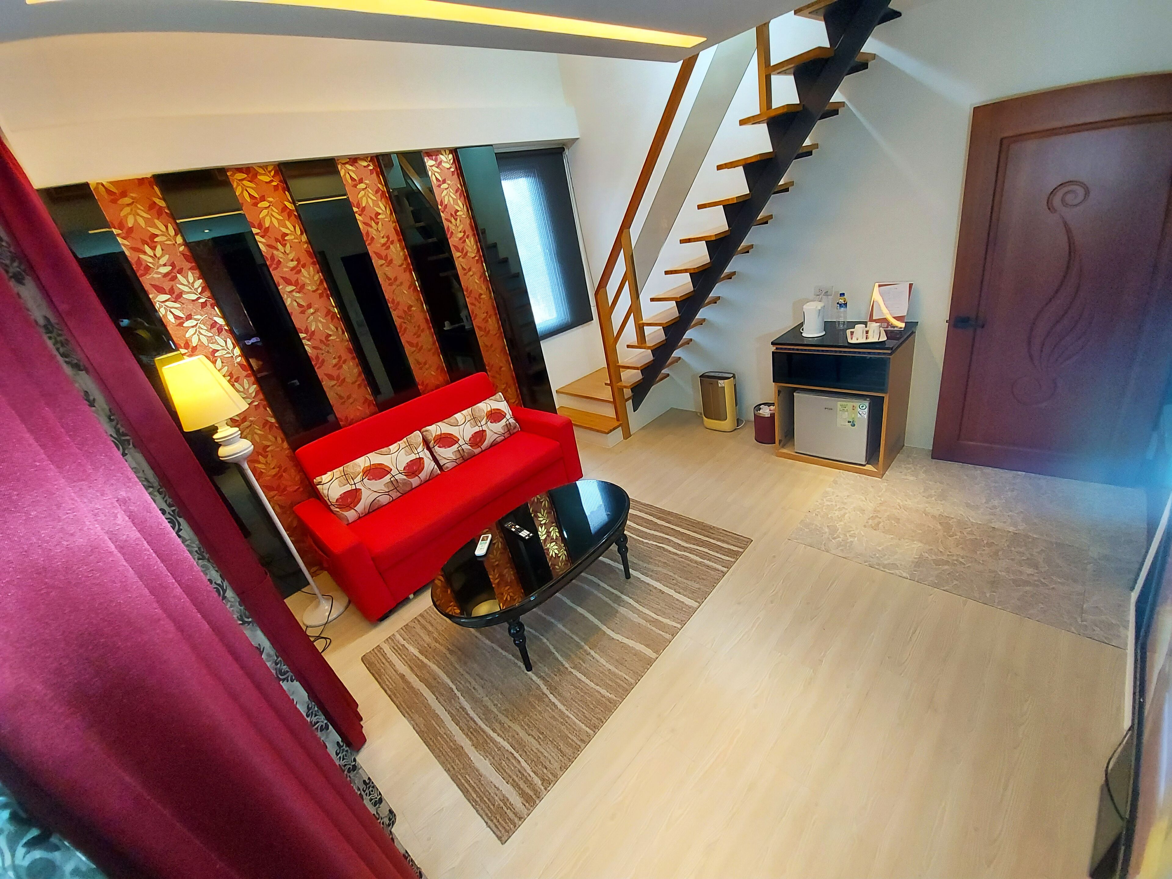 Una sala de estar moderna con un sofá rojo, un sillón negro, una escalera de madera y una pequeña cocina americana.