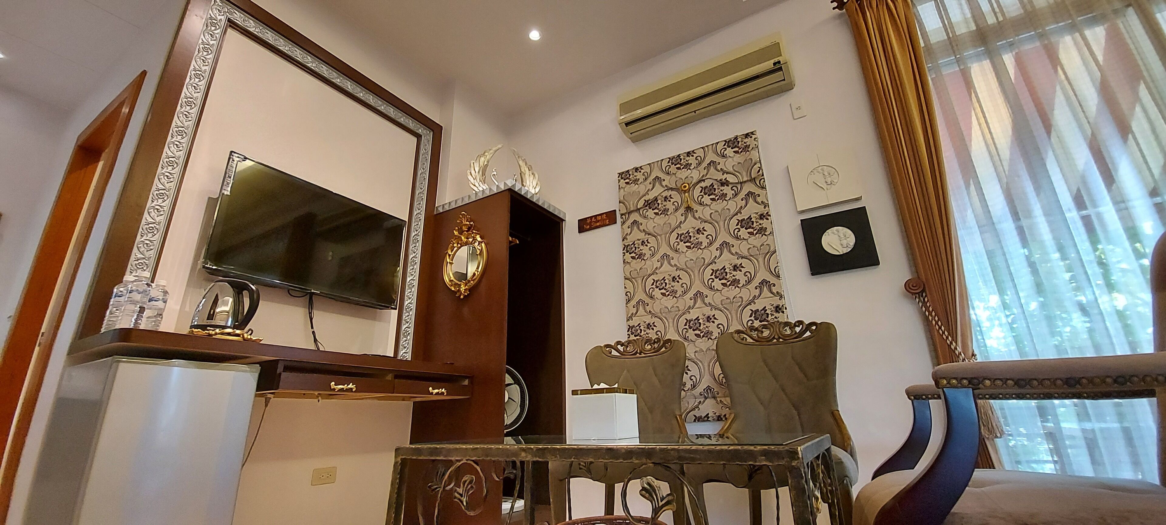 Habitación de hotel con un espejo grande, un televisor de pantalla plana, un mueble de madera, dos sillones tapizados, una mesita y una ventana con cortinas.