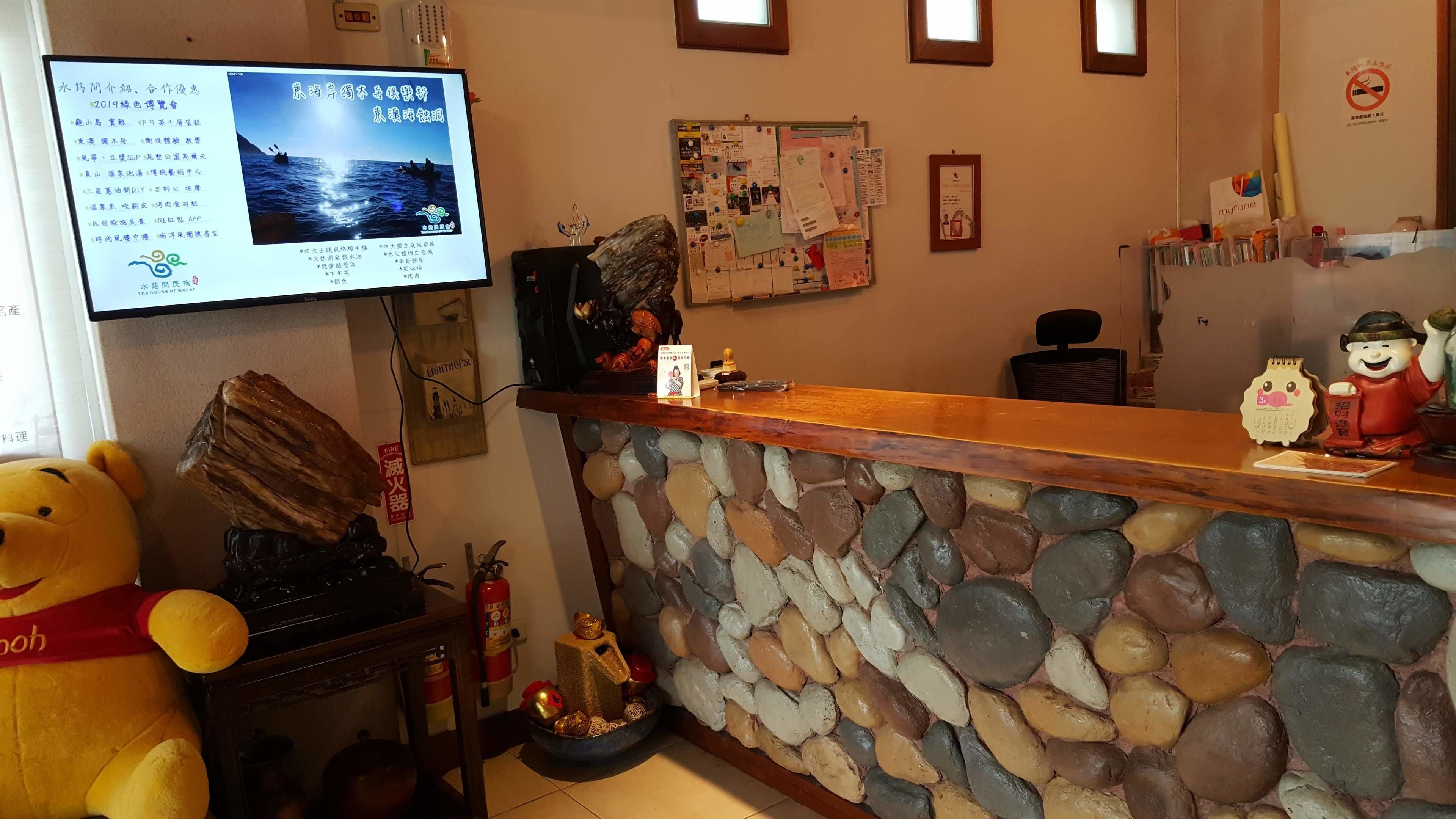 Un área de recepción con pared de piedra, mostrador de madera, televisor mostrando información, un peluche y un extintor.