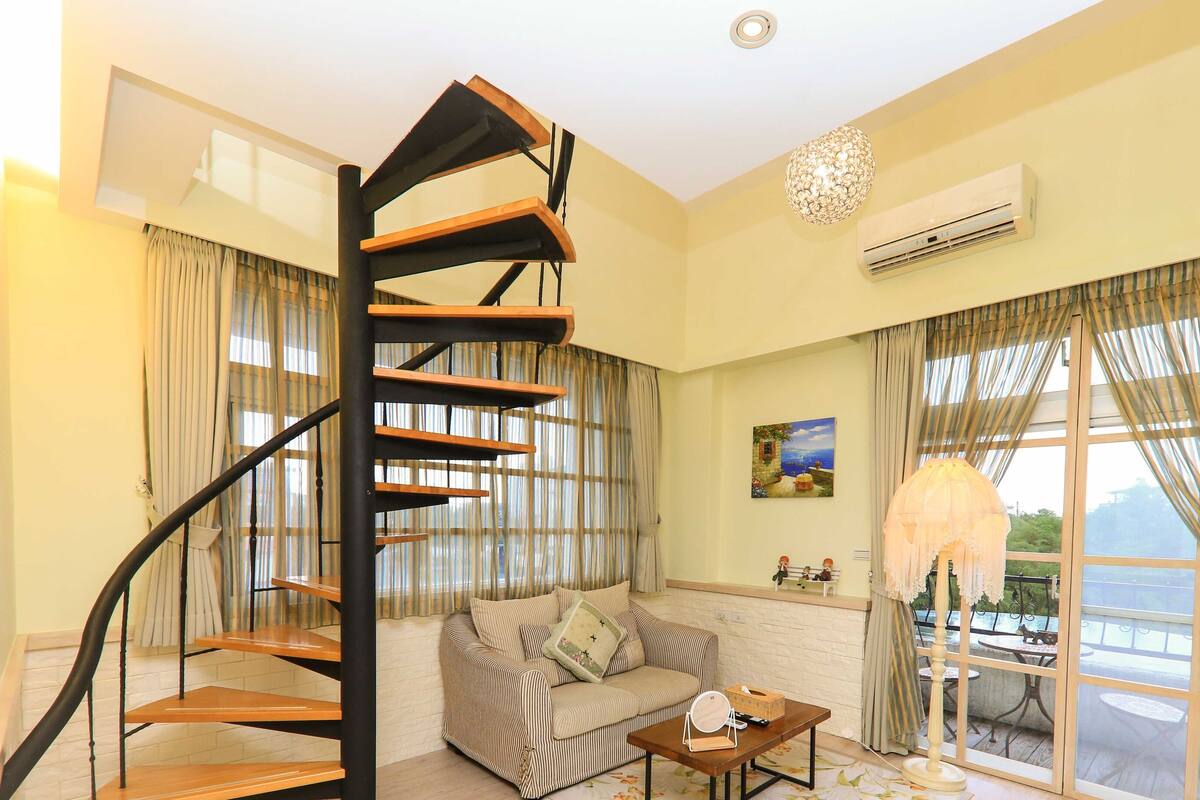 A living room with a spiral staircase, a sofa, a table with items, and a view through large windows.