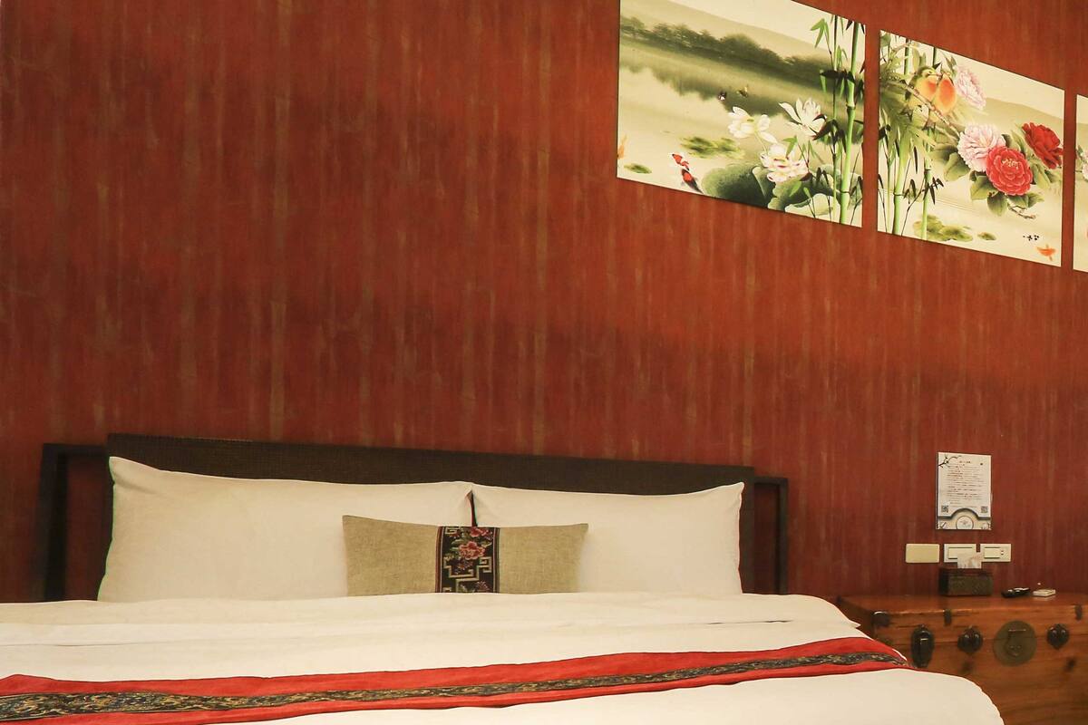 A neatly made bed with white linens and a red decorative border, a wooden headboard, and a wall-mounted painting of flowers.