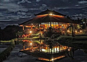 A traditional building with a tiled roof, surrounded by a pond and illuminated by lights. - Puri Mangga Sea View Resort & Spa (Sukasada)