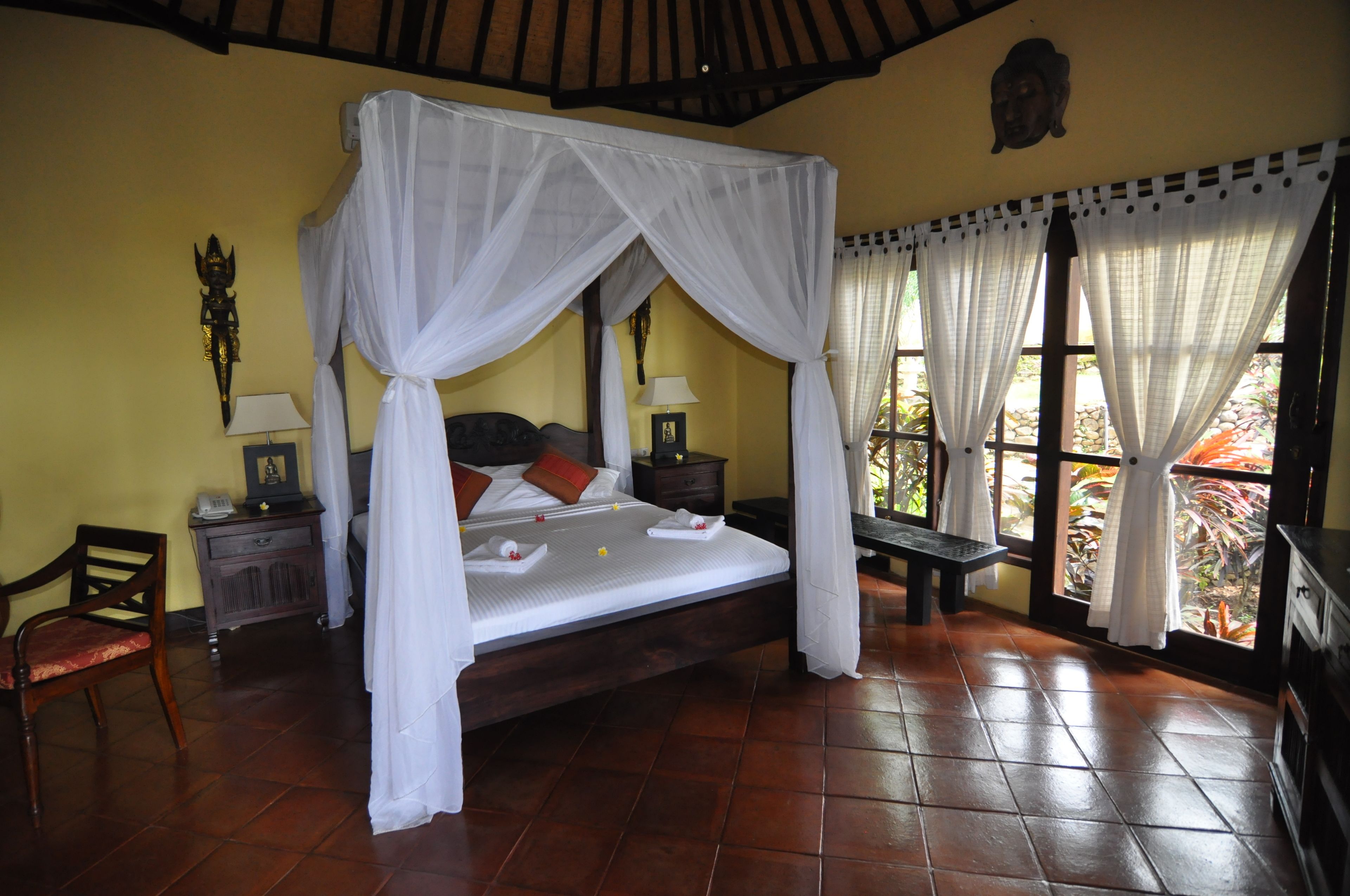 A four-poster bed with white drapes, a wooden nightstand with a lamp, a chair, and a bench.