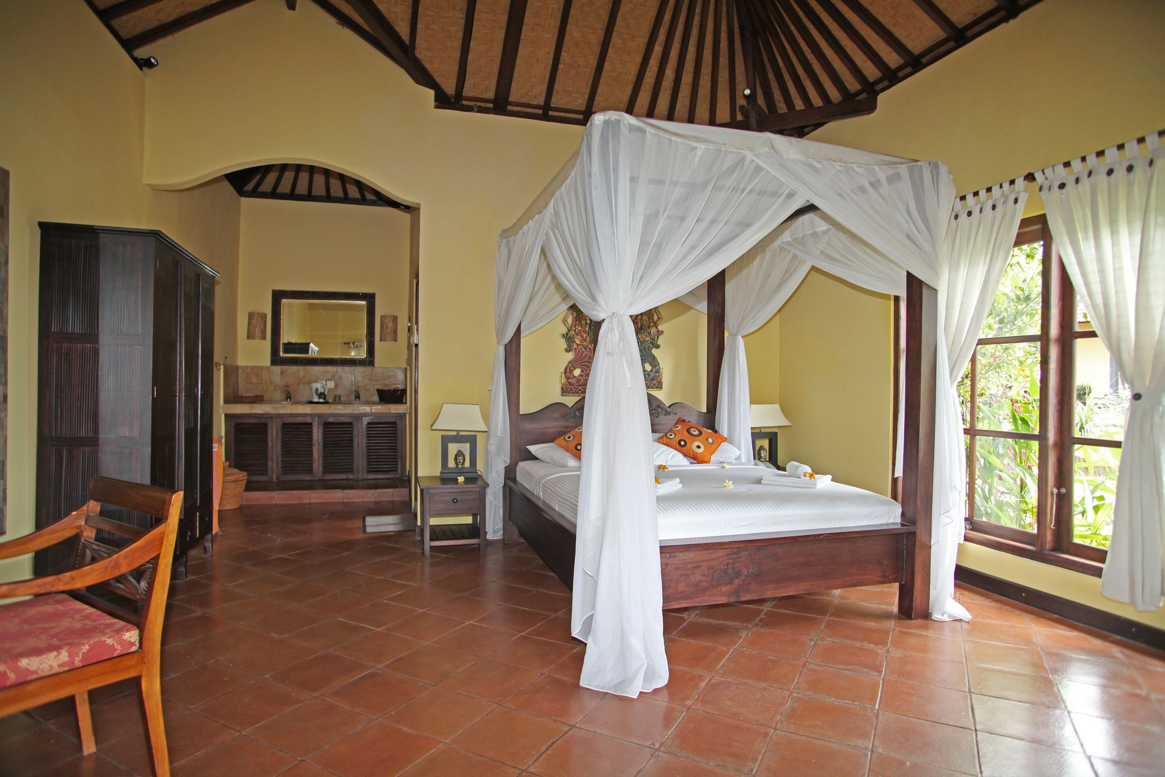 Un dormitorio con cama con dosel, un banco de madera, una tocador con espejo y una ventana con cortinas.