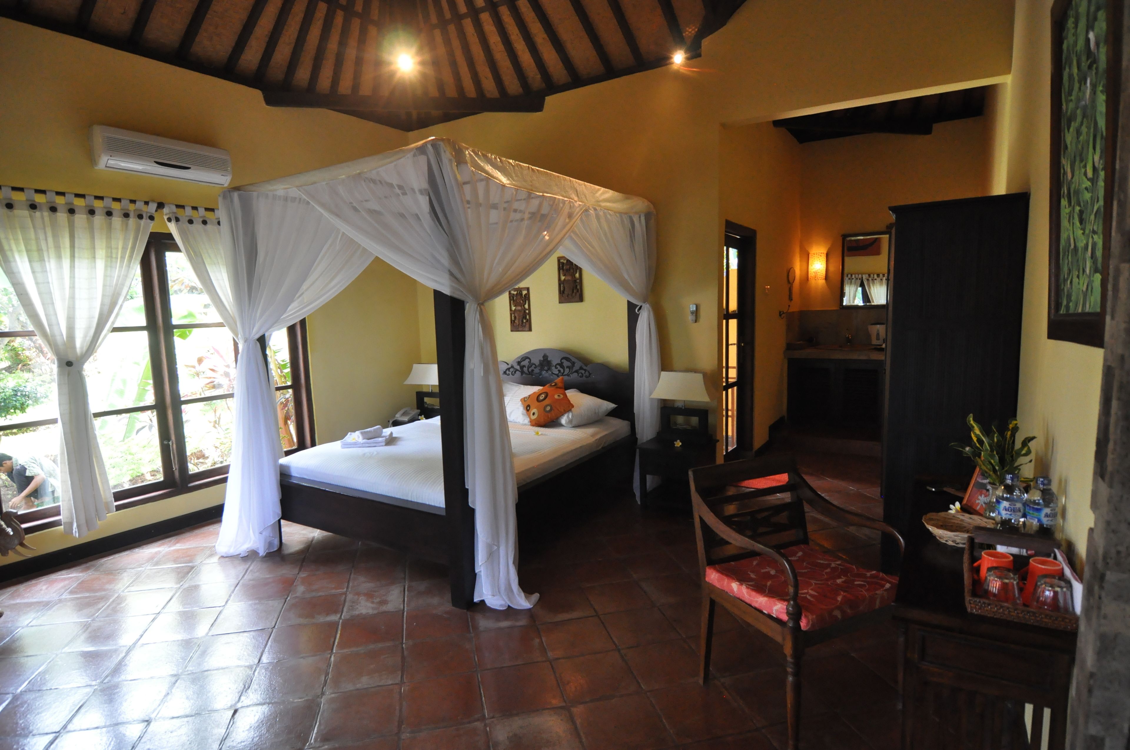 A bedroom with a four-poster bed, a sitting area with a chair, a small table, and a window with curtains.