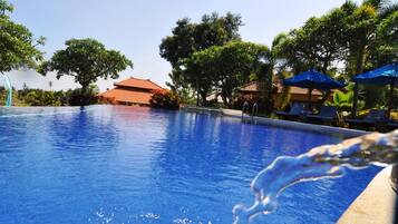 Una piscina azul cristalina con una fuente de agua, rodeada de sillas de descanso y sombrillas, con un edificio al fondo.