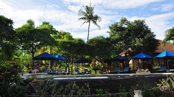 A pool area with lounge chairs and blue umbrellas, surrounded by lush greenery and a palm tree.