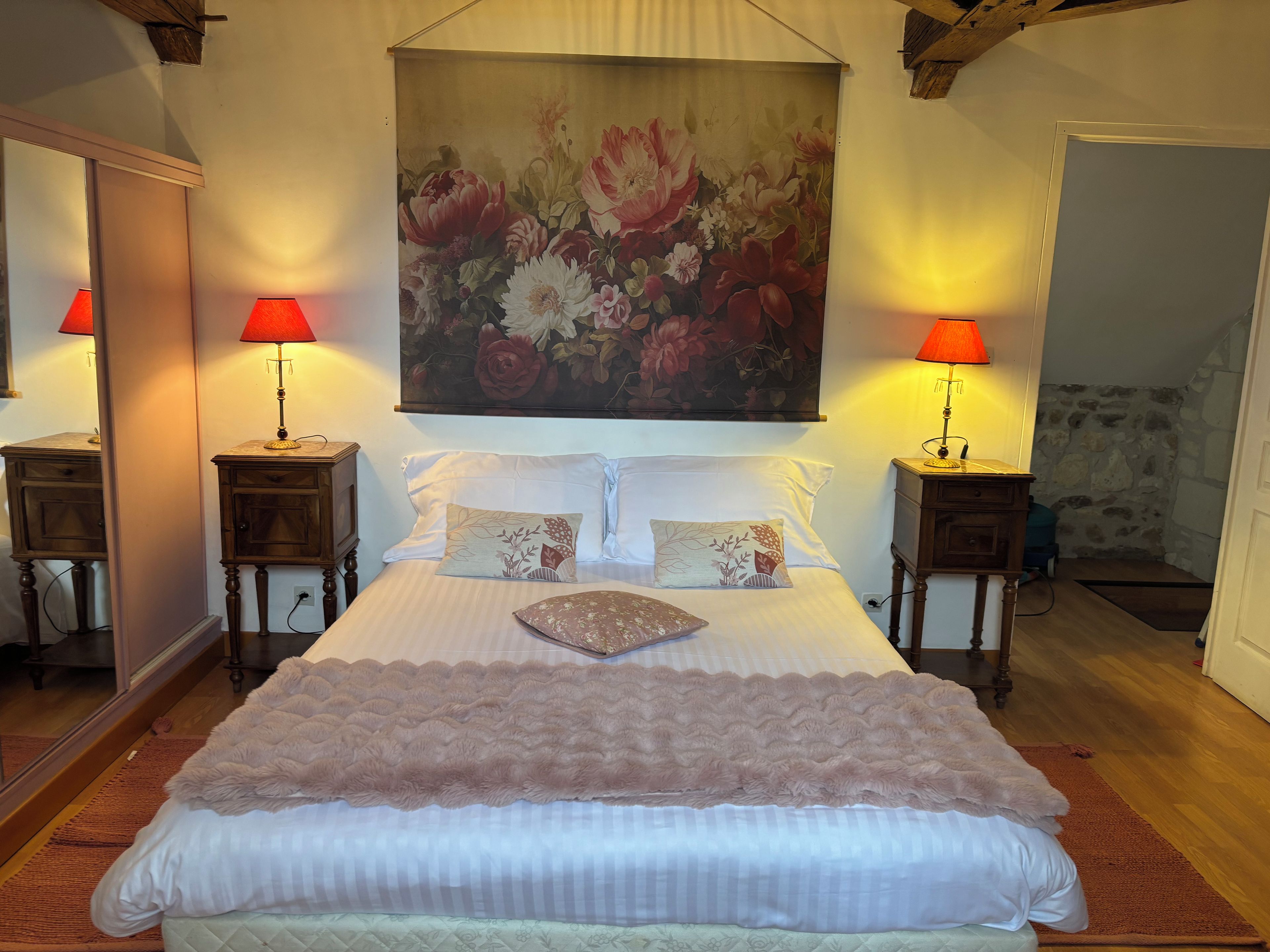 A bedroom with a large bed, two bedside tables with lamps, a floral painting above the bed, and a stone wall in the background.