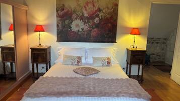 A bedroom with a large bed, two bedside tables with lamps, a floral painting above the bed, and a stone wall in the background.