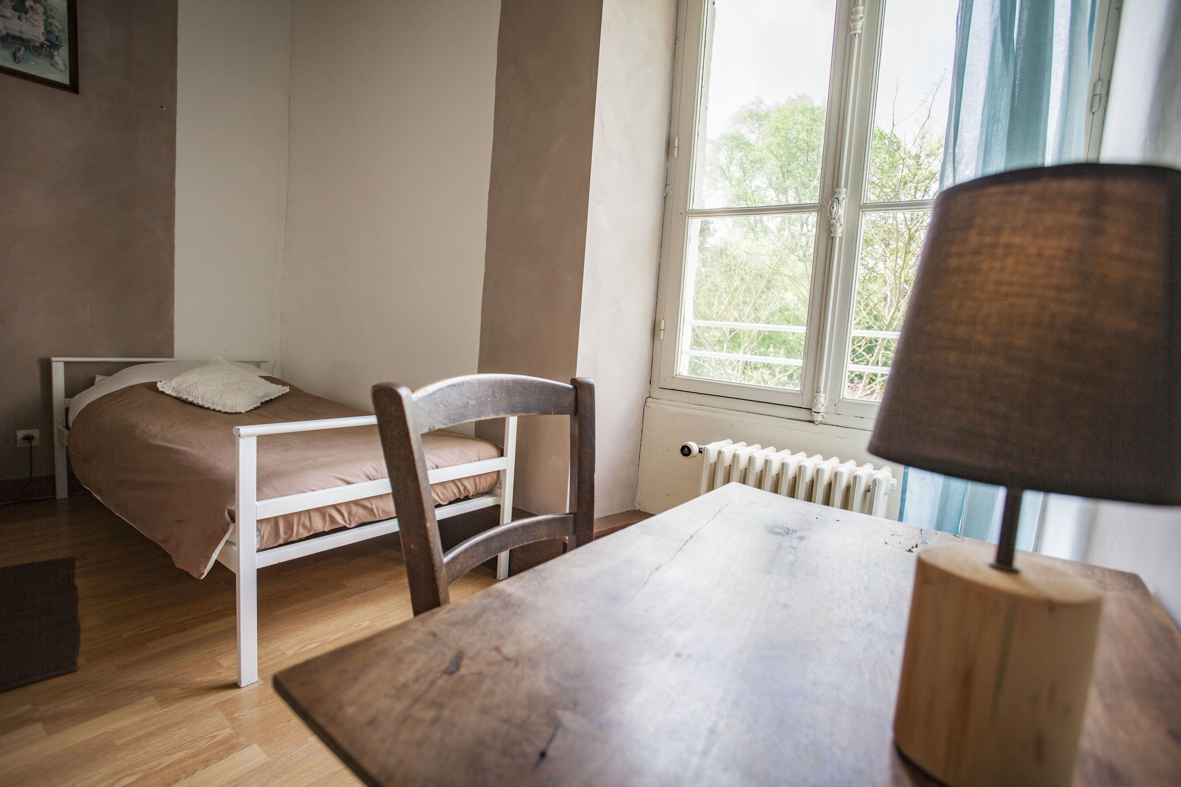 Une chambre à coucher avec un lit, une chaise, une table et une fenêtre donnant sur des arbres.