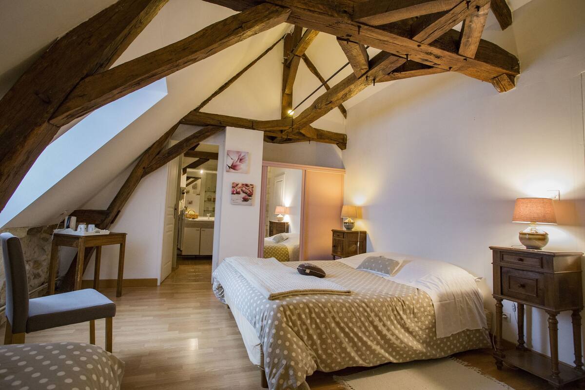 Een slaapkamer met een schuine plafond, houten balken, een bed met een stippenbedekking, een nachtkastje met een lamp, een stoel en een tafeltje met spullen erop.
