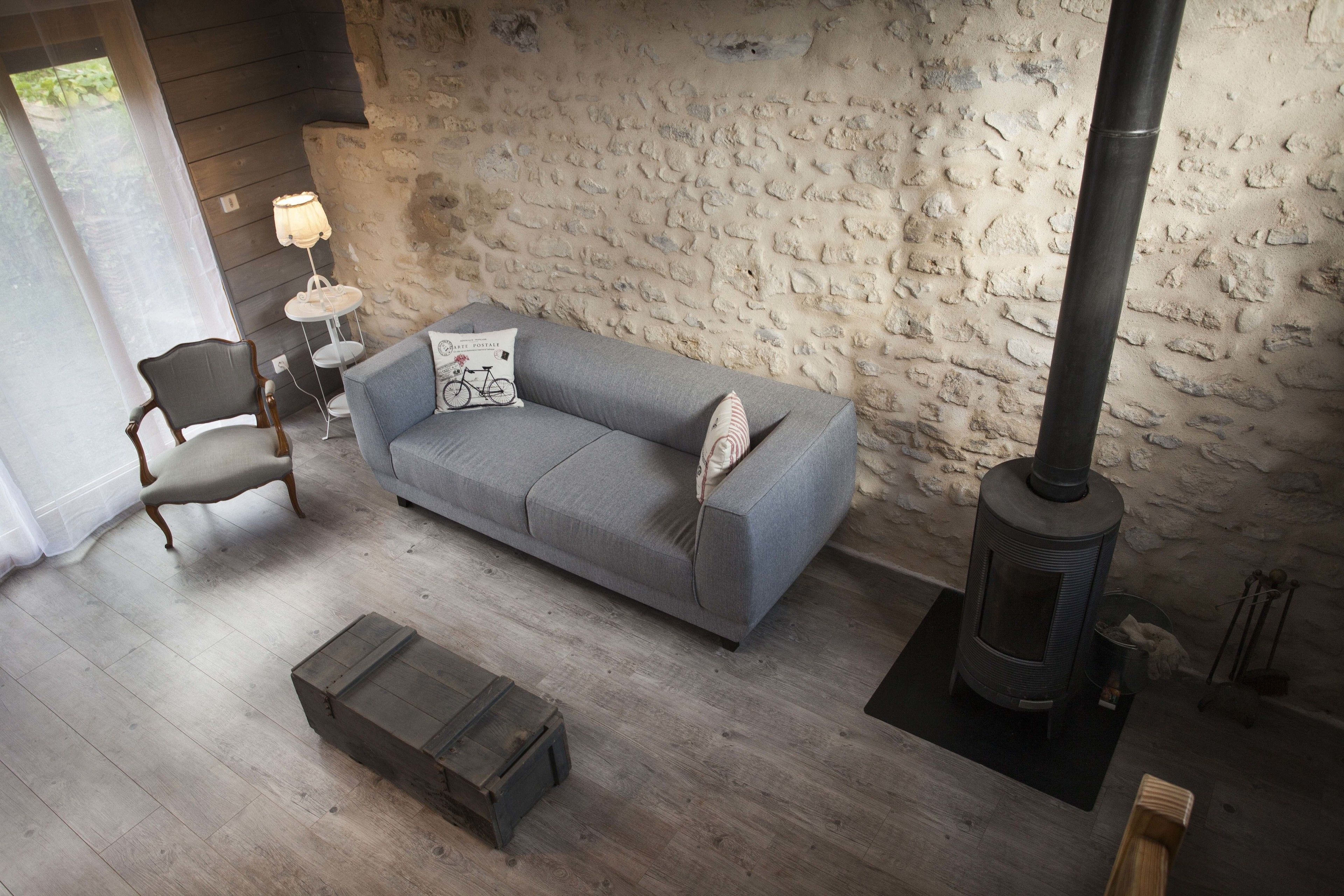 Un salon chaleureux avec un canapé gris, une chaise en bois, une petite table avec une lampe et un poêle à bois adossé à un mur en pierre à la texture intéressante.