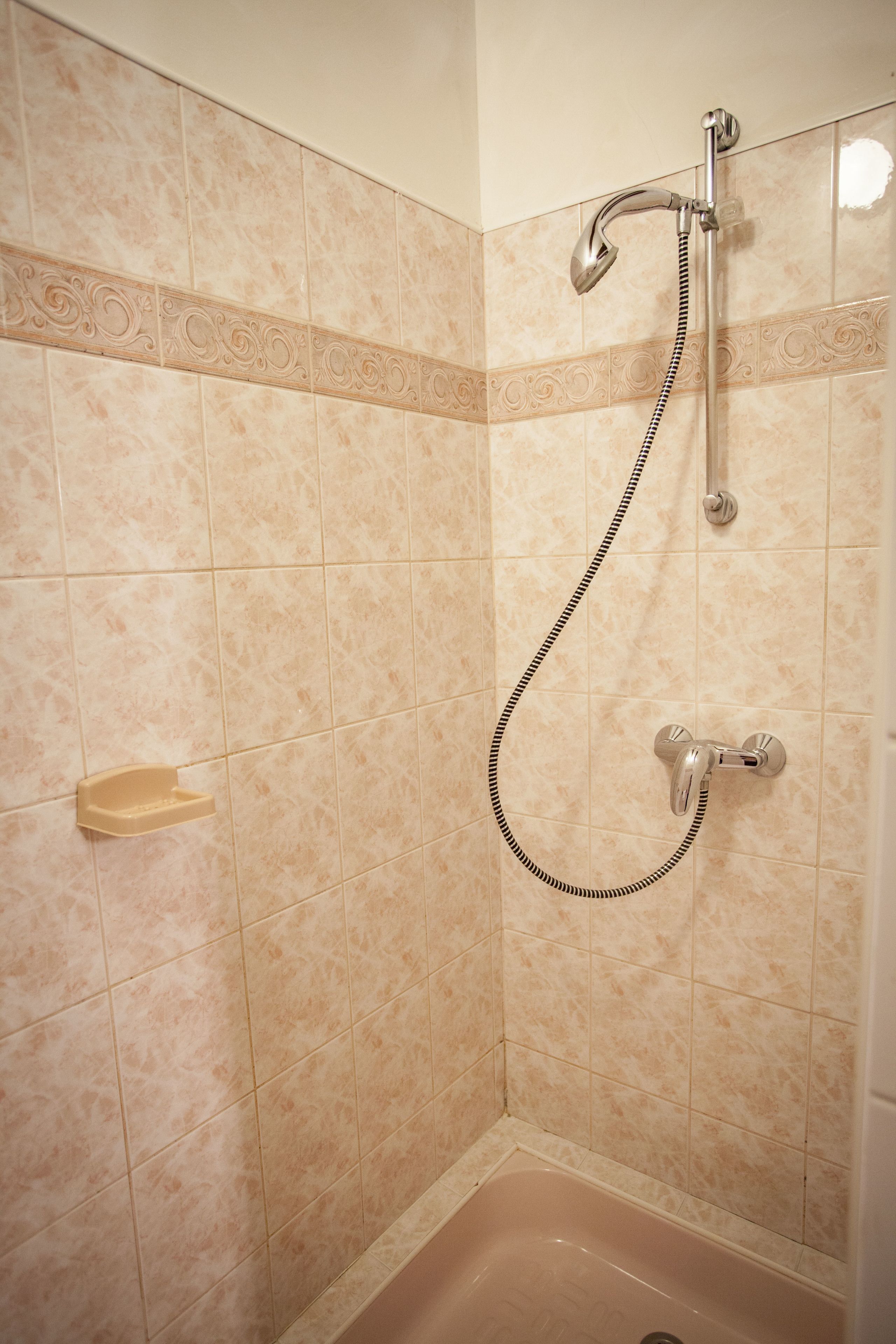 Une salle de bain carrelée de carreaux beiges, une douche avec un pommeau flexible et une petite étagère.