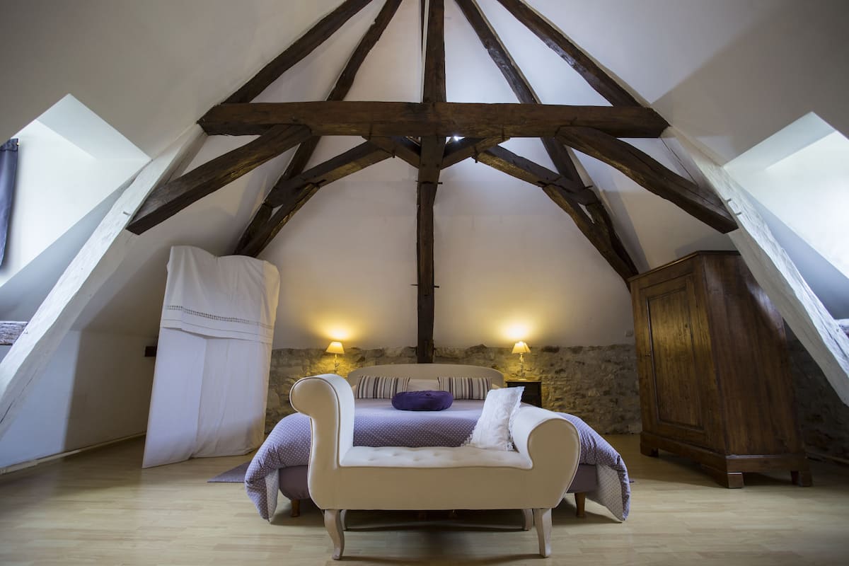 Slaapkamer met schuine plafond, zichtbare houten balken, een bed met een wit hoofdeinde, twee bedlampen en een stenen muur.