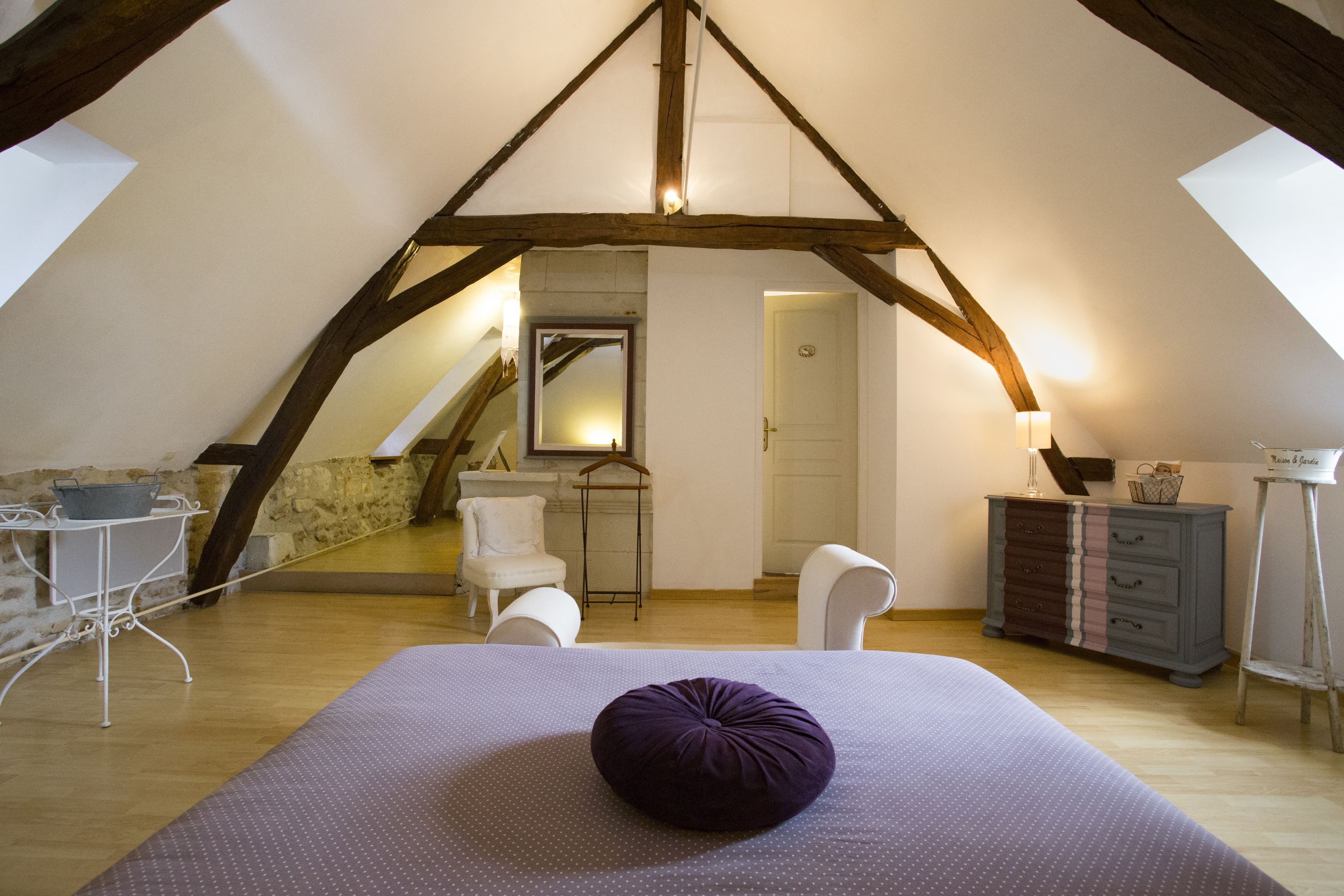 Un dormitorio en el ático con techo inclinado, una cama con almohada morada, un aparador, una silla y una pequeña mesa con una cesta.