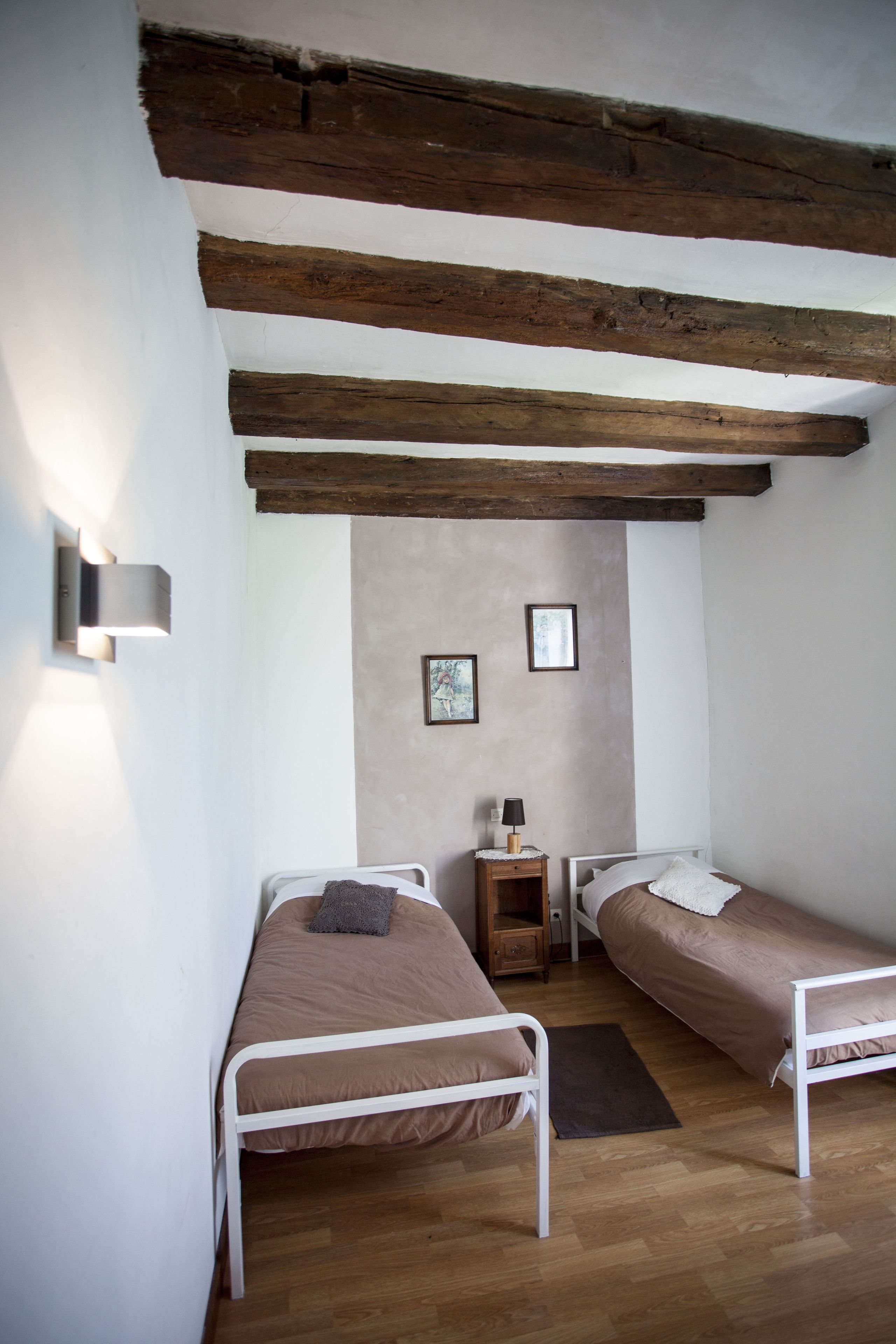 A small, cozy room with two single beds, wooden beams on the ceiling, a small bedside table with a lamp, and a wall-mounted light fixture.
