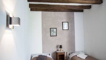 A small, cozy room with two single beds, wooden beams on the ceiling, a small bedside table with a lamp, and a wall-mounted light fixture.