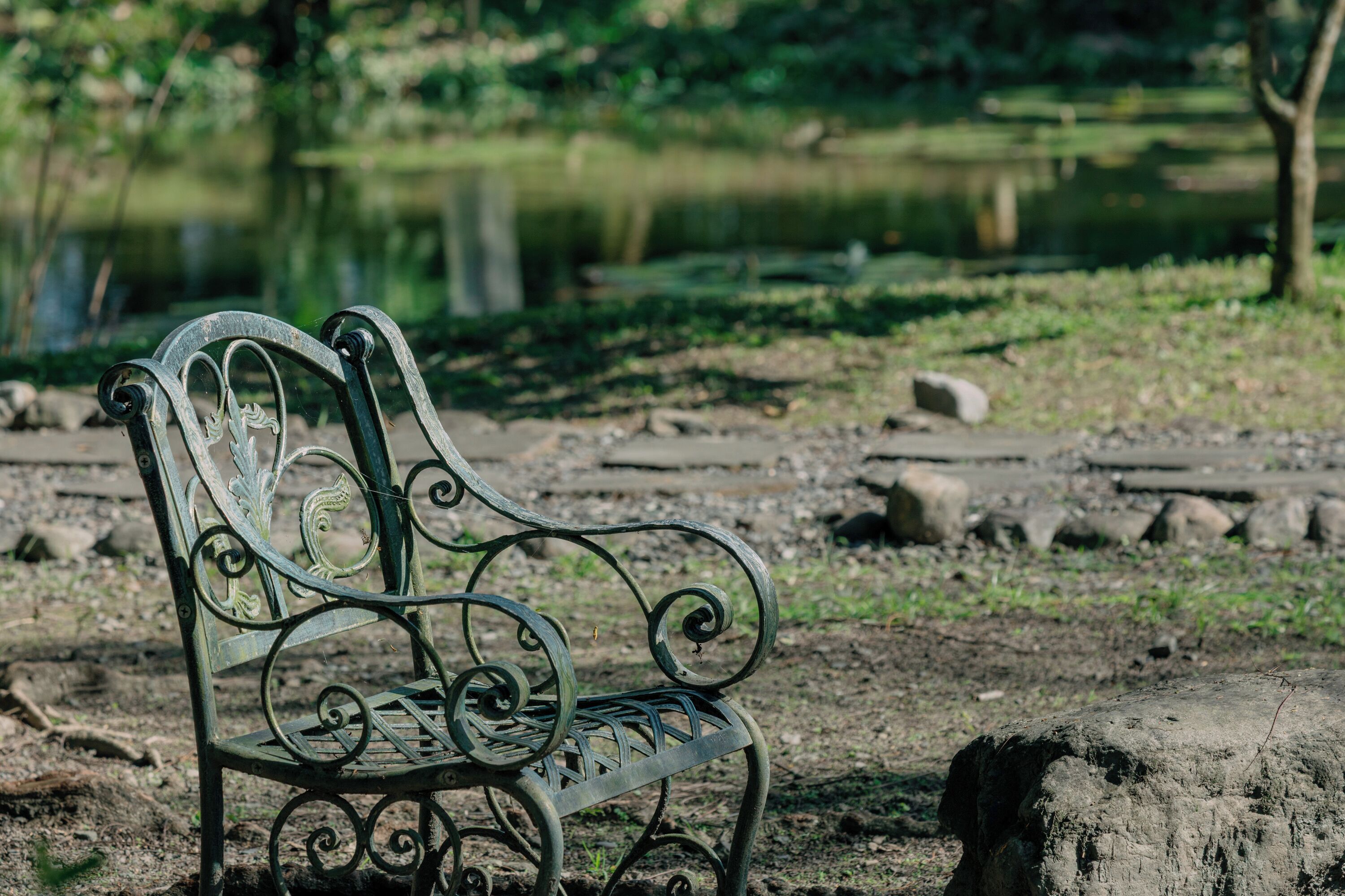 Un banc en métal orné de motifs décoratifs, situé dans un parc avec un étang et une végétation luxuriante en arrière-plan.
