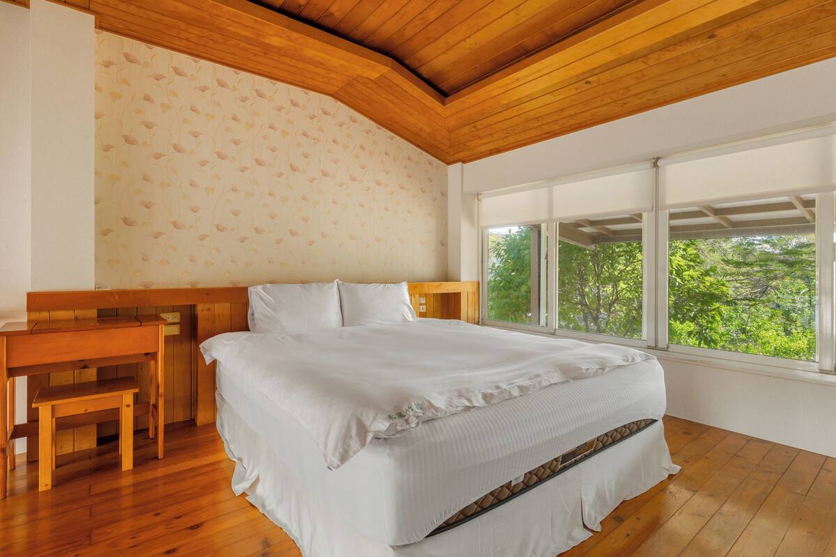 A bedroom with a wooden headboard, a large bed, a wooden bench, and a view of greenery through large windows.