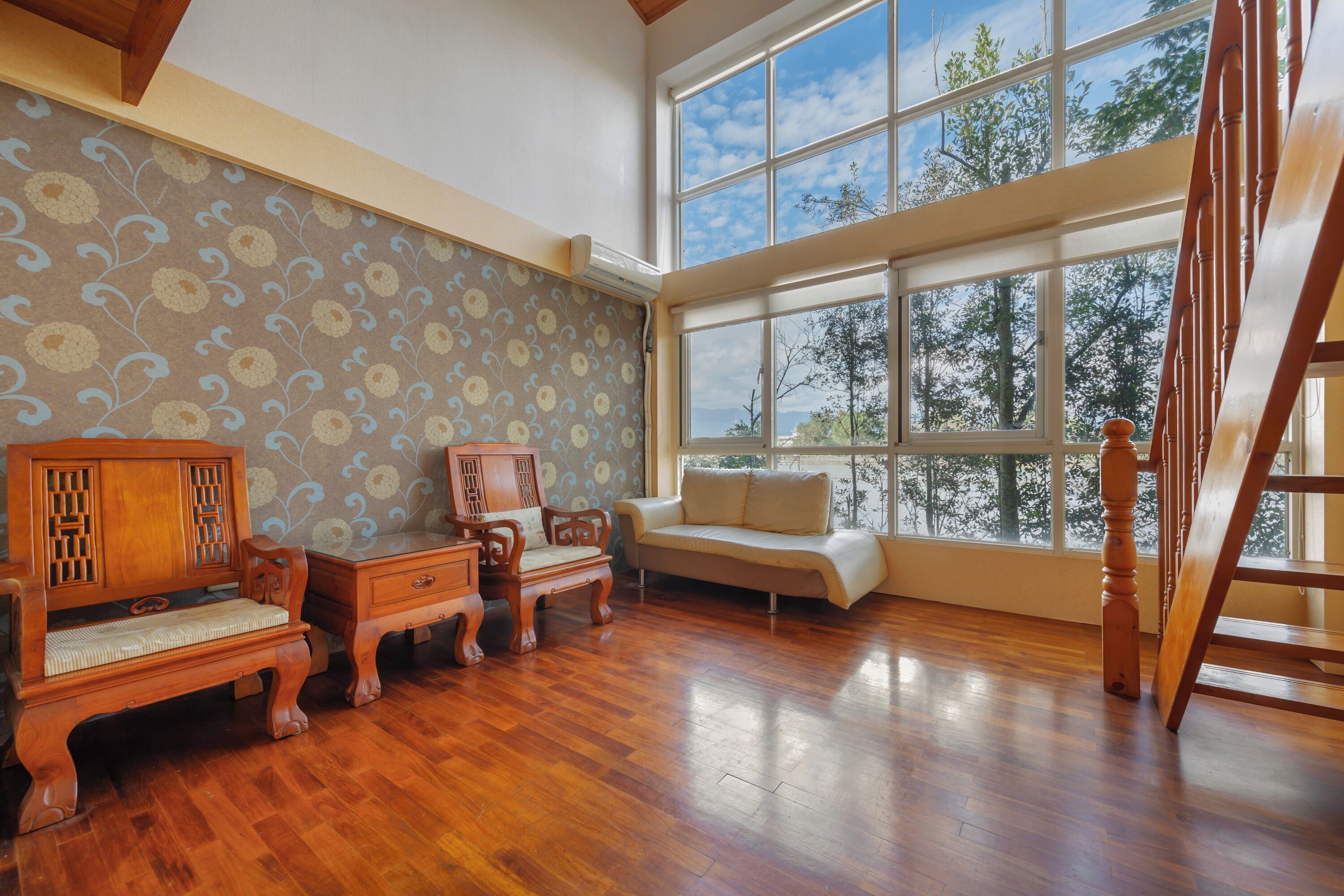 A spacious room with a wooden floor, a sofa, a wooden cabinet, and a large window offering a view of trees.