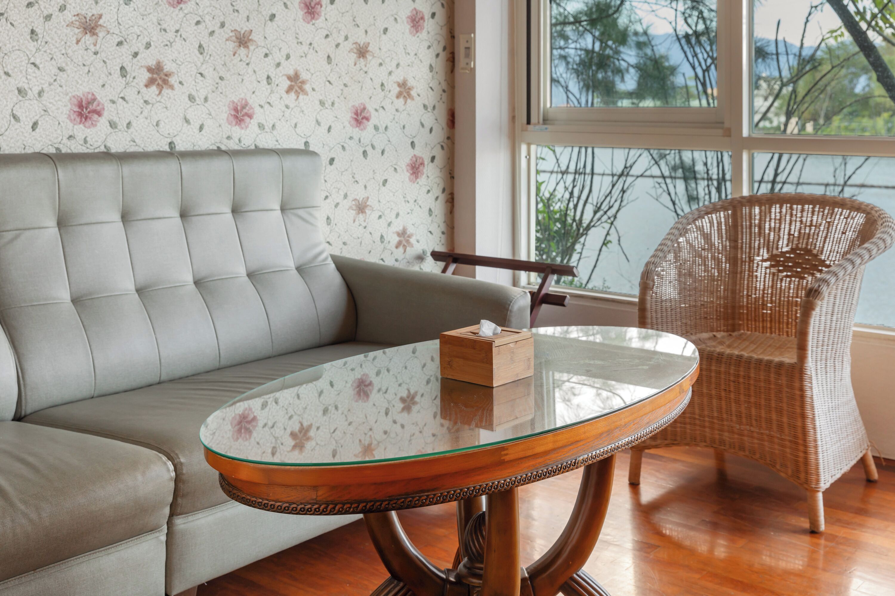Una sala de estar con una mesa de centro de vidrio, un sofá gris, una silla de mimbre y un papel tapiz floral.