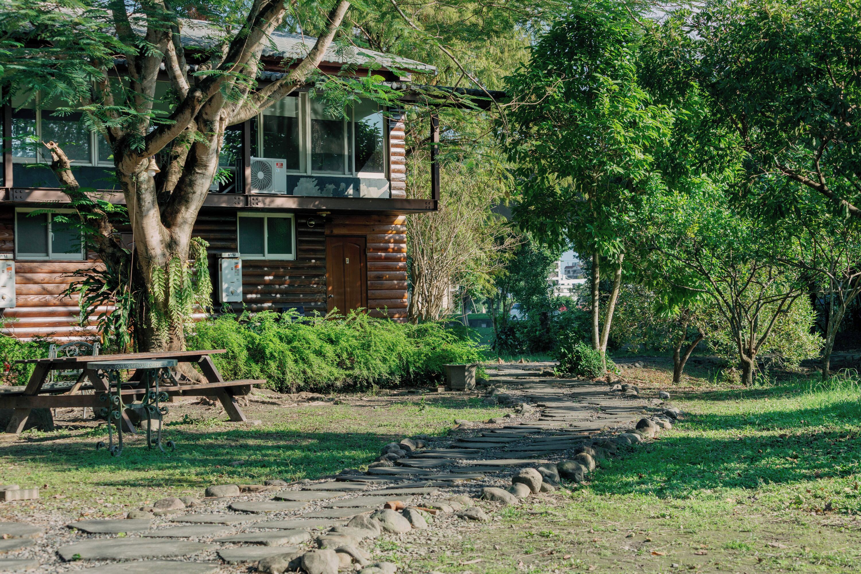 A rustic log cabin with a wooden deck, surrounded by lush greenery and a stone pathway.