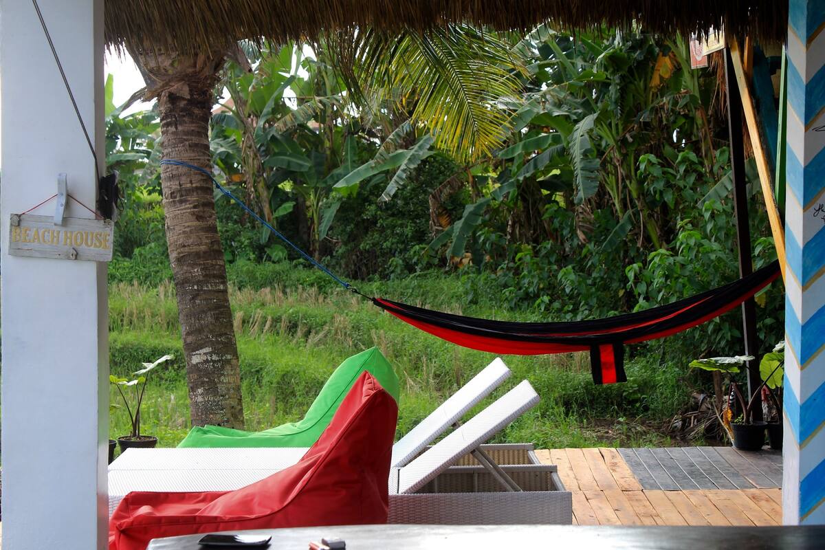 Una hamaca, sillones de descanso y un cartel que dice "BEACH HOUSE" en un entorno tropical.