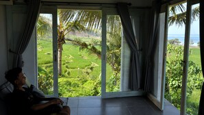 Quarto com uma grande porta de vidro deslizante que dá para uma paisagem verde exuberante, uma pessoa sentada em uma cadeira e vista para o oceano.