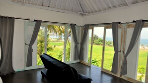 Quarto com sofá preto, janelas amplas com cortinas cinzas e vista para paisagem verde e oceano.