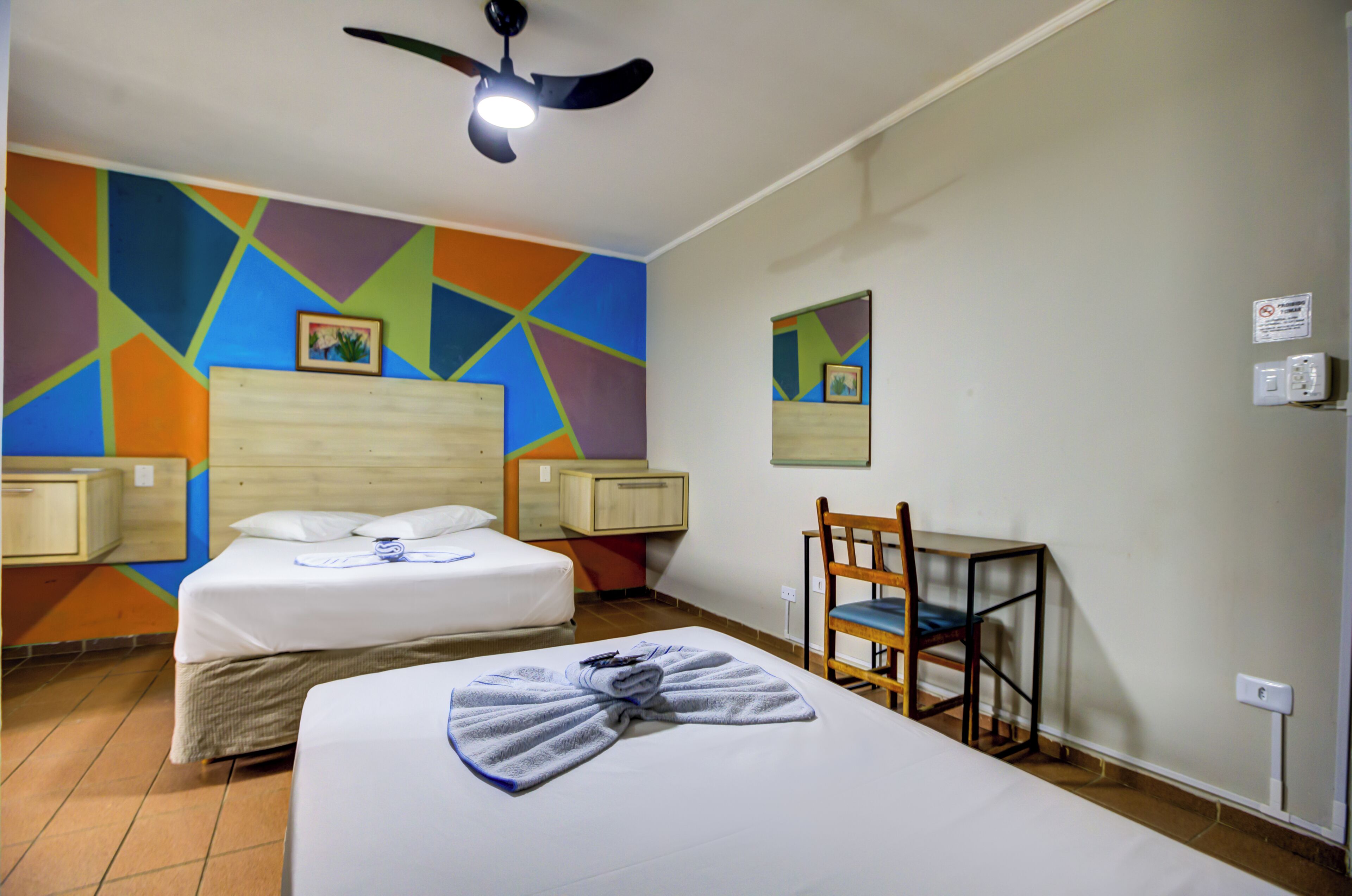 Habitación de hotel con dos camas, ventilador de techo, una silla y arte mural geométrico y colorido.