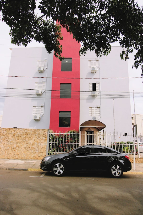 Un edificio de varios pisos con fachada roja y blanca, unidades de aire acondicionado y una entrada protegida por una reja.