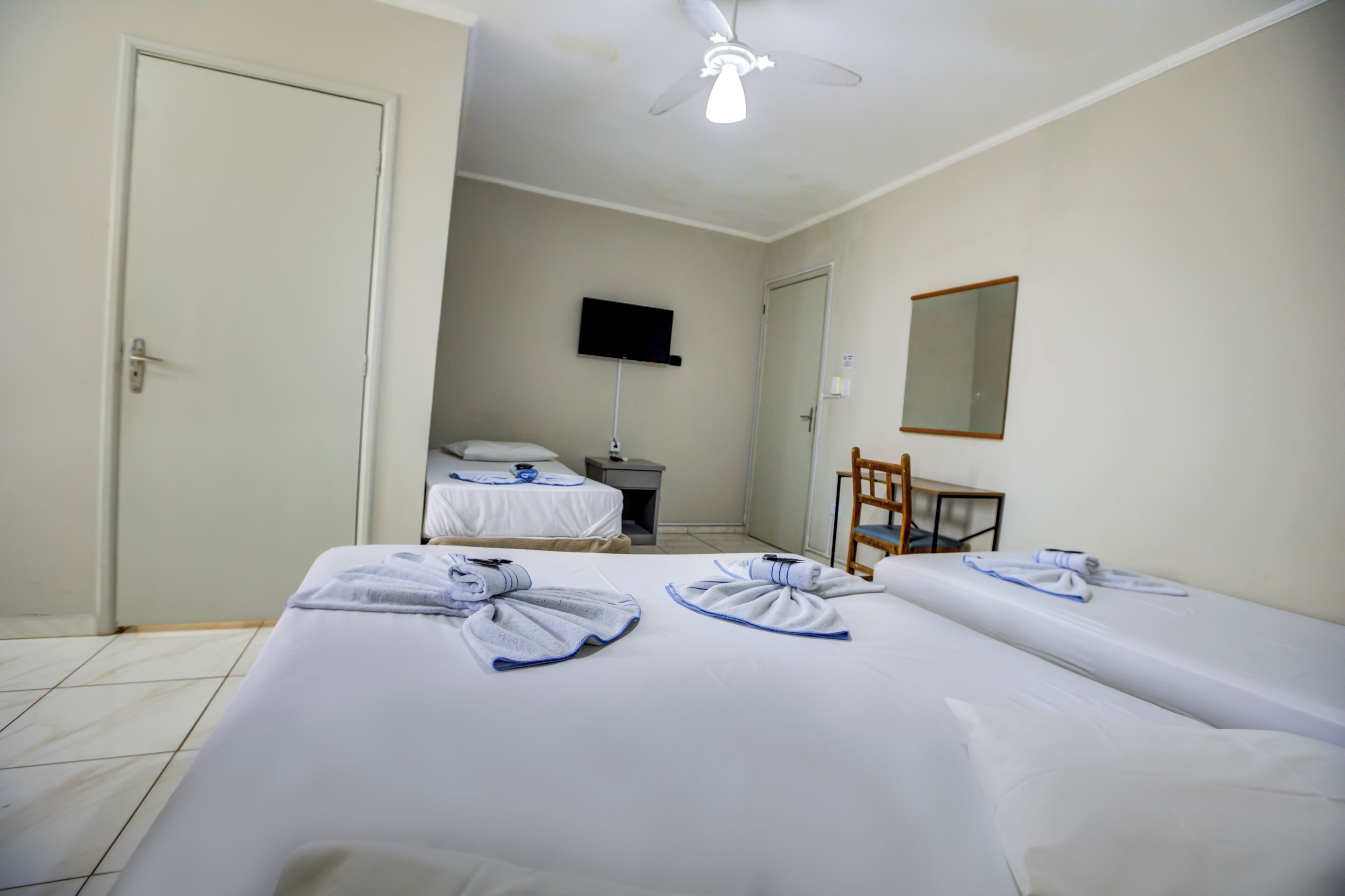 A hotel room with two beds, a TV, a ceiling fan, a chair, and a mirror.