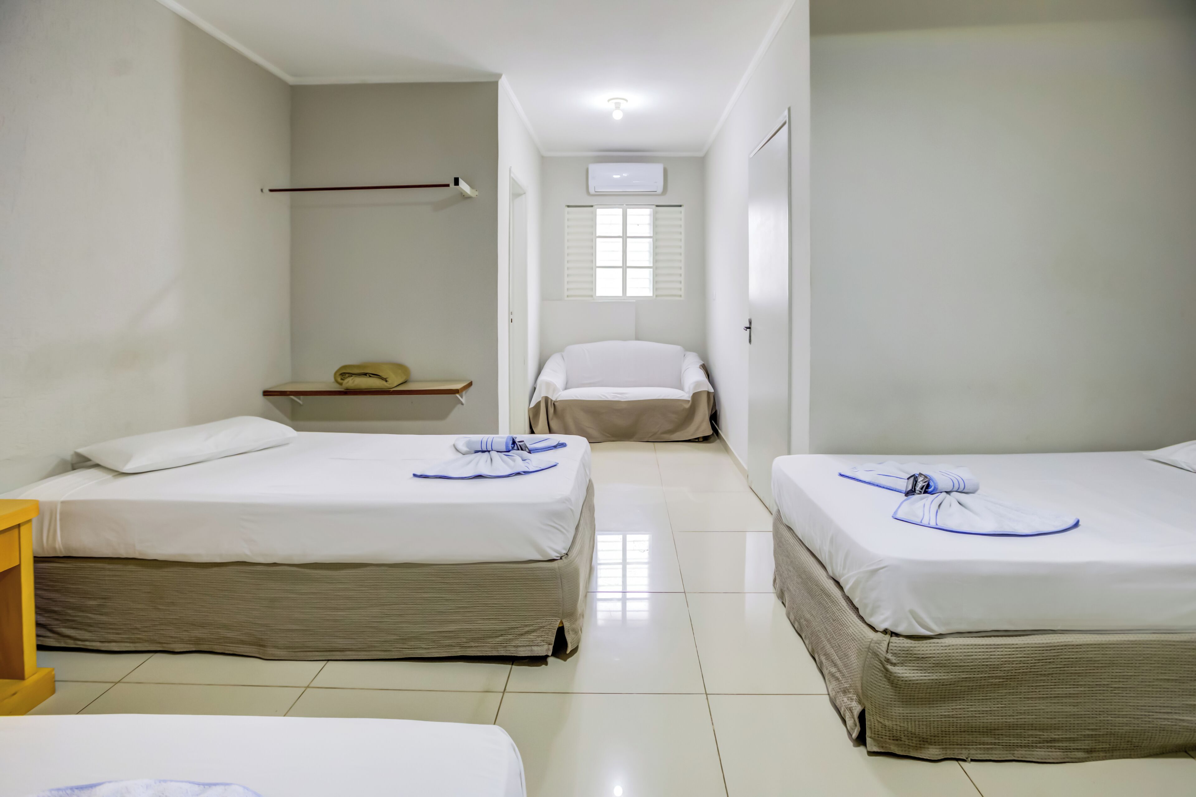 A hotel room with three beds, a small table, a bench, and a window with blinds.