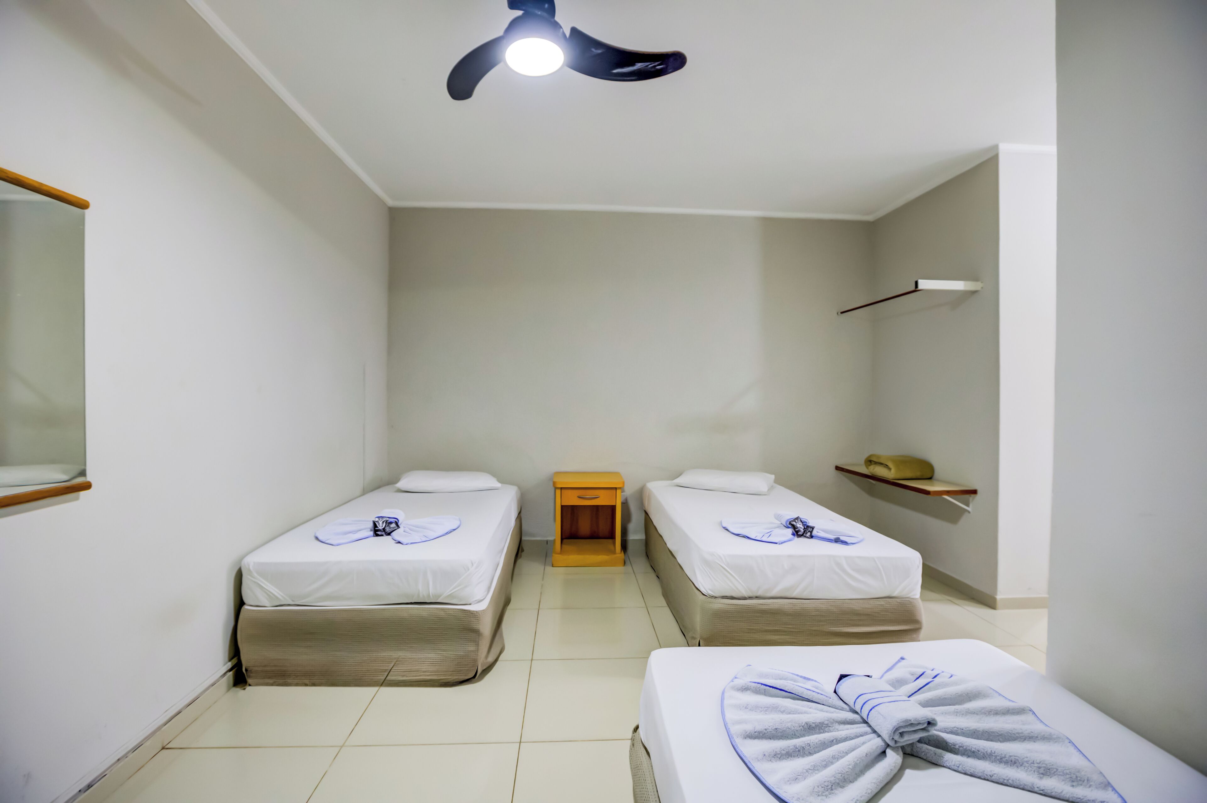 A hotel room with two single beds, a small wooden nightstand, a ceiling fan, and a shelf with a towel.