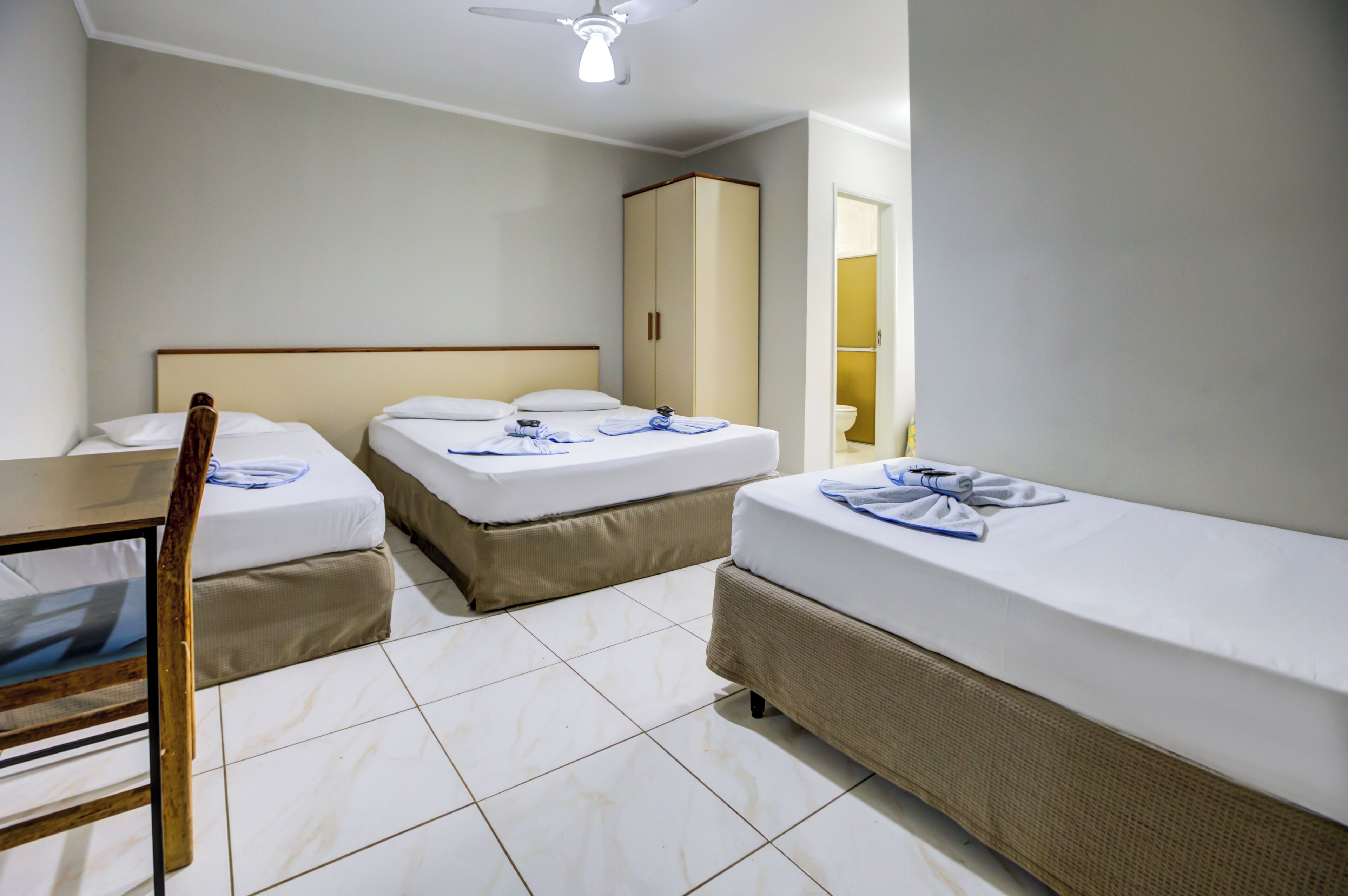 Quarto de hotel com duas camas de solteiro, uma cadeira de madeira, um guarda-roupa e um ventilador de teto.