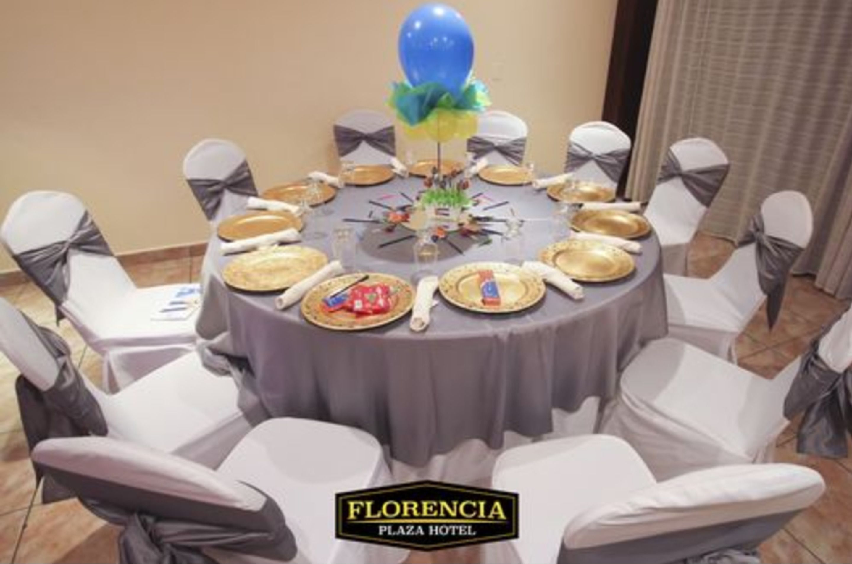 A round table with a grey tablecloth, white chairs with grey sashes, and gold plates.