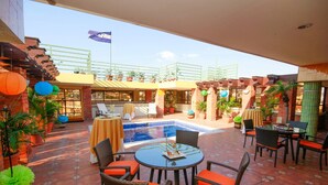 Zona de piscina al aire libre con vistas a la piscina, rodeada de sillas y mesas de mimbre, plantas en macetas y una bandera.