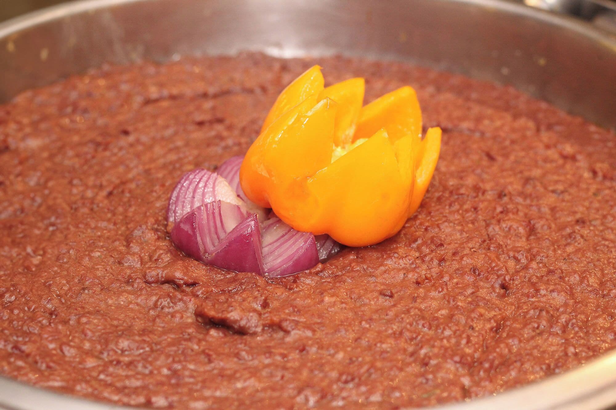 Un tazón de salsa espesa, marrón rojiza, con rodajas de cebolla roja y un pimiento amarillo por encima.