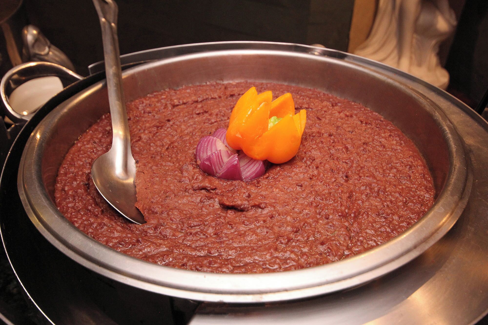 Een grote metalen pan gevuld met een dik, roodbruin gerecht, gegarneerd met rode uien en oranje paprika.