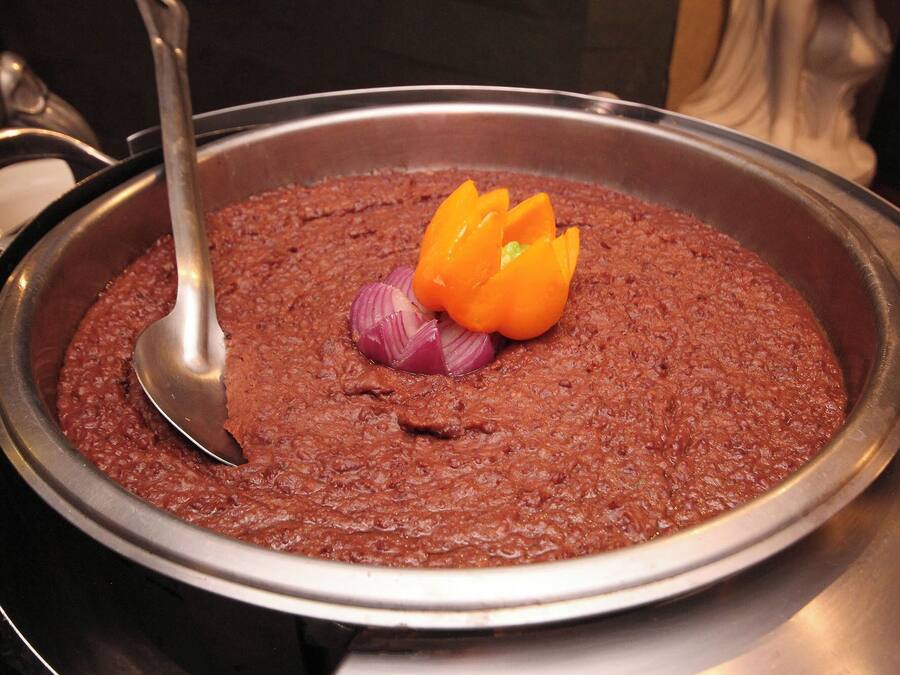 A large metal pan filled with a thick, reddish-brown dish, garnished with sliced red onions and orange bell pepper.
