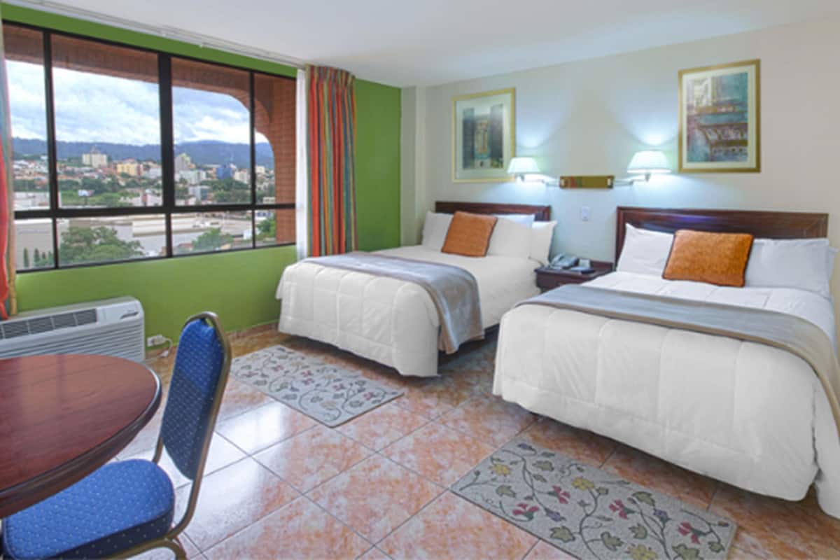 Habitación de hotel con dos camas, una mesa de comedor y vistas a la ciudad a través de amplios ventanales.