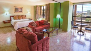 Habitación de hotel con cama, sofá rojo, mesa de centro de vidrio, zona de comedor con mesa y sillas, y vistas a la ciudad a través de amplias ventanas.
