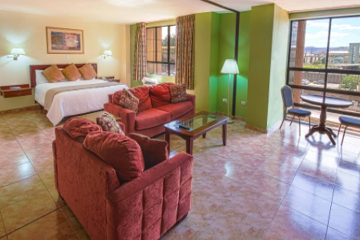 Habitación de hotel con cama, sofá rojo, mesa de centro de vidrio, zona de comedor con mesa y sillas, y vistas a la ciudad a través de amplias ventanas.