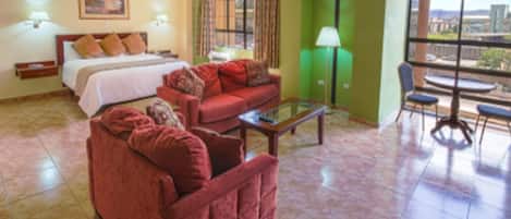 Habitación de hotel con cama, sofá rojo, mesa de centro de vidrio, zona de comedor con mesa y sillas, y vistas a la ciudad a través de amplias ventanas.