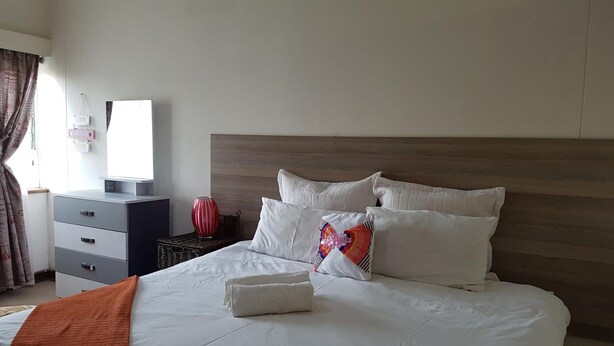 Un lit bien fait, avec une literie blanche et un seul coussin coloré, une table de chevet surmontée d’un vase rouge et un panneau mural portant le mot « VIVRE ».
