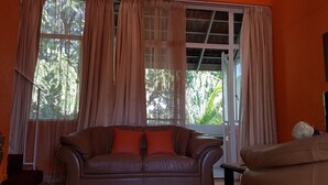 Une pièce avec un canapé en cuir marron, une fenêtre ornée de rideaux légers et une vue sur la verdure à l’extérieur.