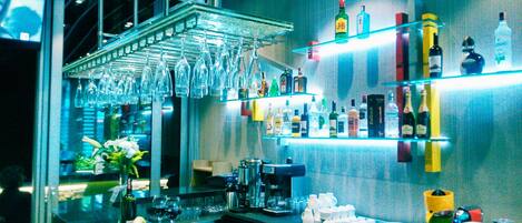 Una zona de bar con mostrador, estantes llenos de botellas, copas colgantes y diversos equipos de bar.