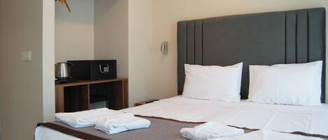 A hotel room with a neatly made bed, a small desk with a kettle, and a coat rack.